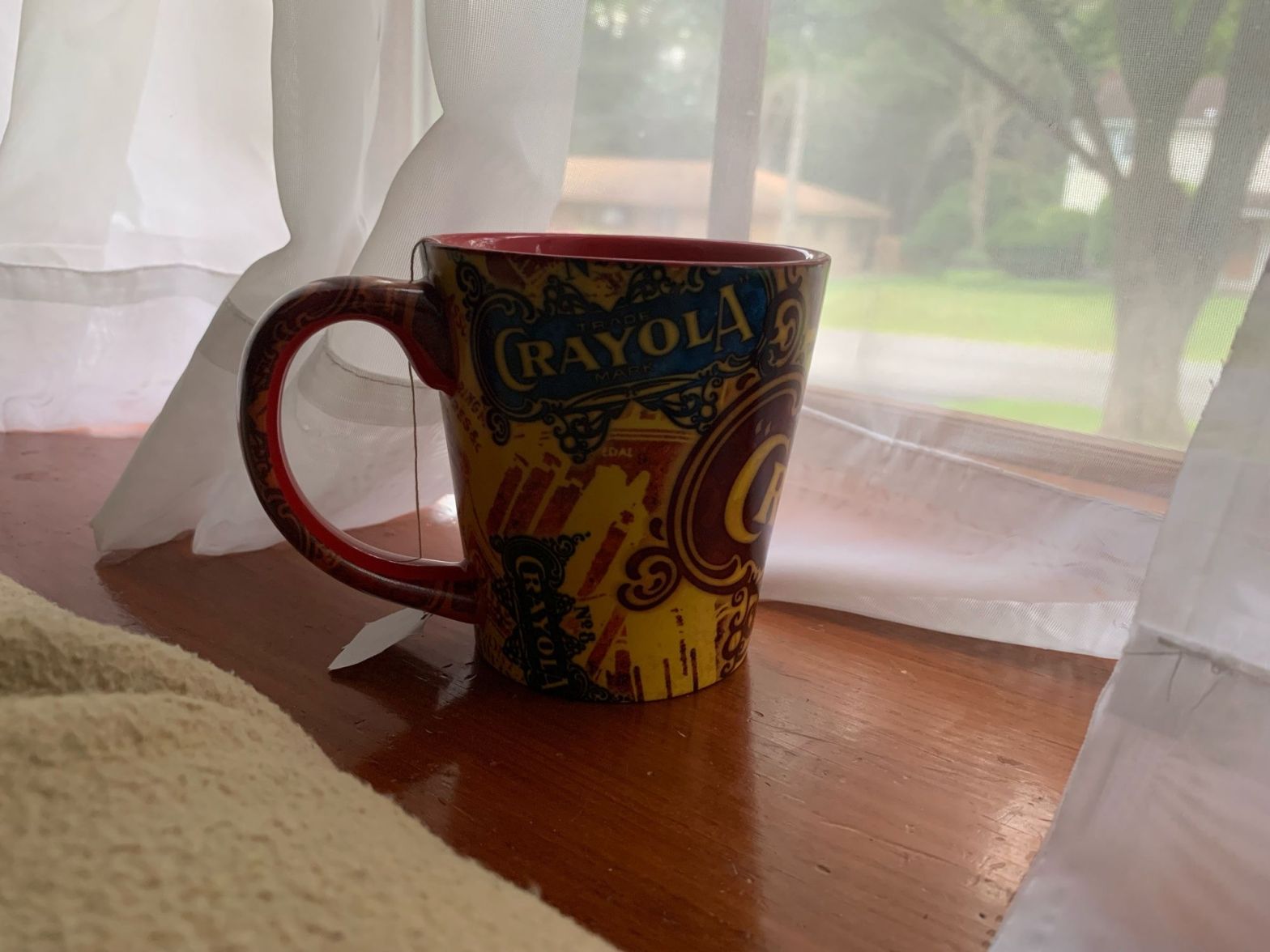 A cup of tea in a Crayola mug sits on a windowsill.